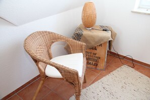 Una silla de mimbre con un cojín blanco, un jarrón tejido sobre una mesita y un saco de arpillera con texto en el suelo.