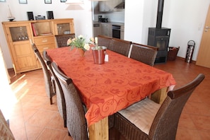 Un coin repas avec une table dressée pour six personnes, entourée de chaises en osier. Un buffet en bois avec des portes en verre se trouve à gauche, et on aperçoit un espace cuisine au fond.