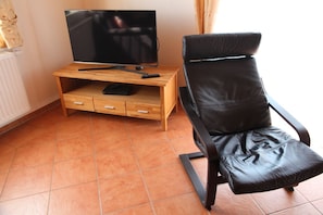 Un salon avec un téléviseur posé sur un meuble en bois, un fauteuil en cuir et un sol carrelé.