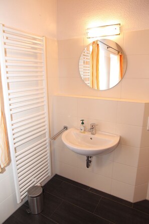 Une salle de bain avec un lavabo blanc, un miroir rond, un porte-serviettes et une poubelle.