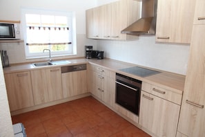A modern kitchen with light wood cabinets, a stainless steel range hood, a built-in oven, and a window with curtains.