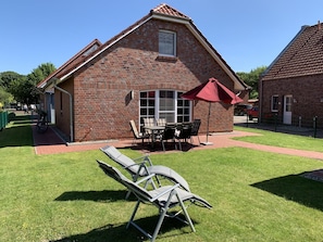 A brick building with a sloped roof, a patio with a table and chairs, and a lawn with lounge chairs. - Aloha - Aloha in Greetsiel: 3 bedrooms, wood-burning stove, garden and WLAN (Krummhörn)