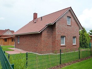 A single-story brick house with a red-tiled roof, a visible chimney, and a green metal fence in the foreground. - Aloha - Aloha in Greetsiel: 3 bedrooms, wood-burning stove, garden and WLAN (Krummhörn)