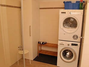 A compact laundry area with a washing machine and dryer, a small stool, and a shelf. - Aloha - Aloha in Greetsiel: 3 bedrooms, wood-burning stove, garden, and Wi-Fi (Krummhörn)