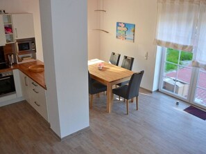 A modern kitchen with a dining area, wooden table, chairs, and a view through a glass door. - Aloha - Aloha in Greetsiel: 3 bedrooms, wood-burning stove, garden, and Wi-Fi (Krummhörn)