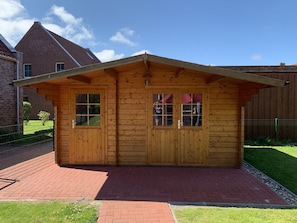 A wooden cabin with a pitched roof, two windows, and a brick patio. - Aloha - Aloha in Greetsiel: 3 bedrooms, wood-burning stove, garden and WLAN (Krummhörn)