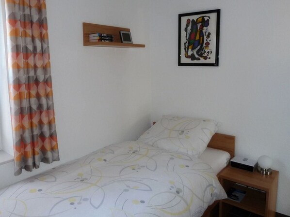 A small, neatly arranged bedroom with a single bed, a bedside table with a lamp, a shelf with books and a phone, and a colorful abstract painting on the wall.