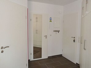 Un pasillo con paredes blancas, piso de baldosas y puertas que dan a habitaciones. Las puertas tienen manijas y hay una pequeña señal en la pared.