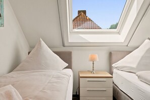 Ein Schlafzimmer mit Oberlicht, zwei Betten, einem Nachttisch mit Lampe und Blick auf ein Gebäude durch das Fenster.