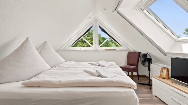 A bedroom with a bed, pillows, a chair, a fan, a TV, and a skylight.