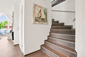 Una escalera de madera conduce a una plataforma, con un cuadro en la pared y vistas a un área exterior con piscina.