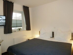 Une chambre d’hôtel avec un lit, une fenêtre avec des rideaux, un radiateur, une table de chevet et un téléviseur fixé au mur.