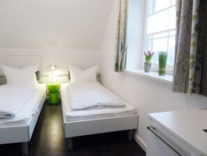 Une chambre d’hôtel avec deux lits simples, une table de chevet avec une lampe, une petite fenêtre avec des rideaux et une plante en pot sur le rebord de la fenêtre.