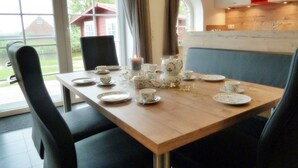 A dining table set with plates, cups, and a candle, surrounded by chairs in a room with large windows and a view of a house. - Riedhaus am Siel - 100sqm vacation home for 6 pers. + dog with fireplace, heat cabin & (Krummhörn)