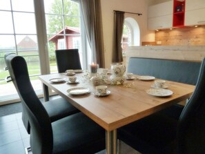 A dining table set with plates, cups, and a candle, surrounded by chairs in a room with large windows and a view of a house.
