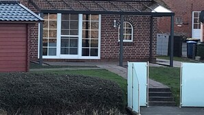 A brick house with a covered porch, a small garden, and a visible trash bin. - Riedhaus am Siel - 100sqm vacation home for 6 pers. + dog with fireplace, heat cabin & (Krummhörn)
