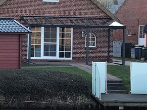 Una casa de ladrillo con un porche cubierto, un pequeño jardín y un bote de basura visible.