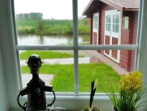 A window with a view of a red building and a pond. - Riedhaus am Siel - 100sqm vacation home for 6 pers. + dog with fireplace, heat cabin & (Krummhörn)