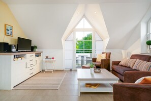 Een moderne woonkamer met een witte bank, een salontafel en een televisie op een witte kast. Een groot raam biedt uitzicht op het landschap.