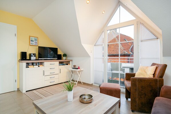 Een moderne woonkamer met een hoge nok, een televisie op een houten plank, een witte kast, een klein tafeltje met een plant en een bruine leren bank.