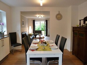 Uma área de refeições com uma mesa branca posta para quatro pessoas, rodeada por cadeiras, e vista para o exterior através de uma porta de vidro.