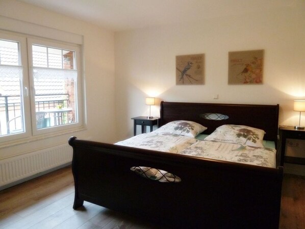 A bedroom with a wooden bed, two wall-mounted lamps, a window with a view, and two framed pictures on the wall.