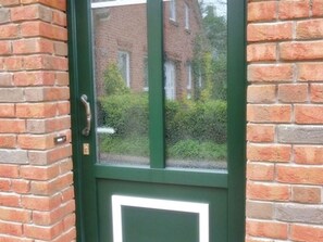 Uma porta verde com uma janela retangular, paredes de tijolo e uma maçaneta visível.