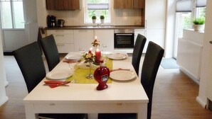 Une cuisine moderne avec des meubles blancs, une table à manger pour quatre personnes et une fenêtre équipée de stores.