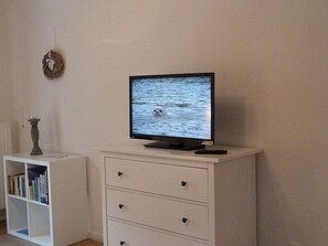 A television on a white cabinet, a white bookshelf, and a wall-mounted decorative item. - Gesine's Huuske - Gemütliches Ferienhaus im Eilsumer Ortskern (Krummhörn)