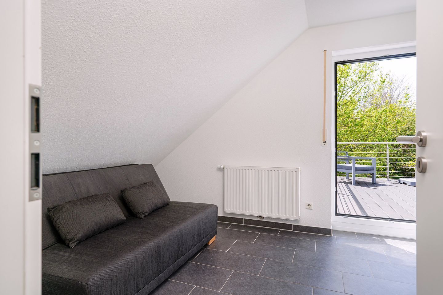 Et moderne loftværelse med en grå sofa, en hvid radiator og udsigt til en balkon med gelænder og grønt.