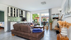 Una sala moderna con un sofá marrón, una mesita de centro y una zona de comedor con una mesa y sillas de madera. Se observa una cocina con armarios blancos y un salpicadero verde. Hay una planta en maceta y un florero con flores secas sobre una mesita de madera.