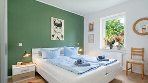 Un dormitorio con una cama, dos mesitas de noche, una lámpara auxiliar, una ventana con vistas, una silla y obras de arte en las paredes.