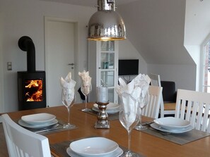 Mesa de comedor dispuesta con platos blancos, cubiertos y cristalería. Arriba, una lámpara colgante moderna, y en el fondo, una estufa negra con fuego visible.