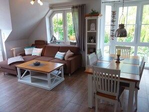 Un salón acogedor con un sofá marrón, una mesa de centro de madera y una mesa de comedor con sillas. Hay una estantería con objetos decorativos y una ventana con cortinas.