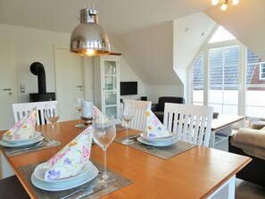 Ein Esstisch ist gedeckt mit Tellern, Gläsern und Servietten, darüber hängt eine Chandelier und durch ein Fenster ist ein Haus zu sehen.