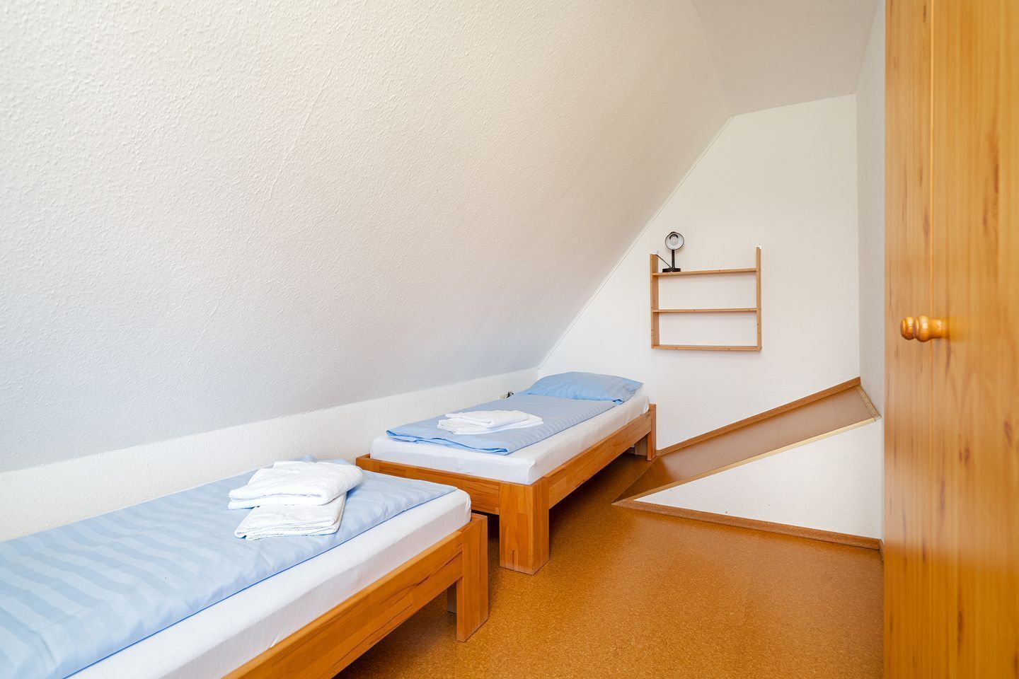 A small, compact room with two single beds, a wooden door, and a small shelf with a lamp.