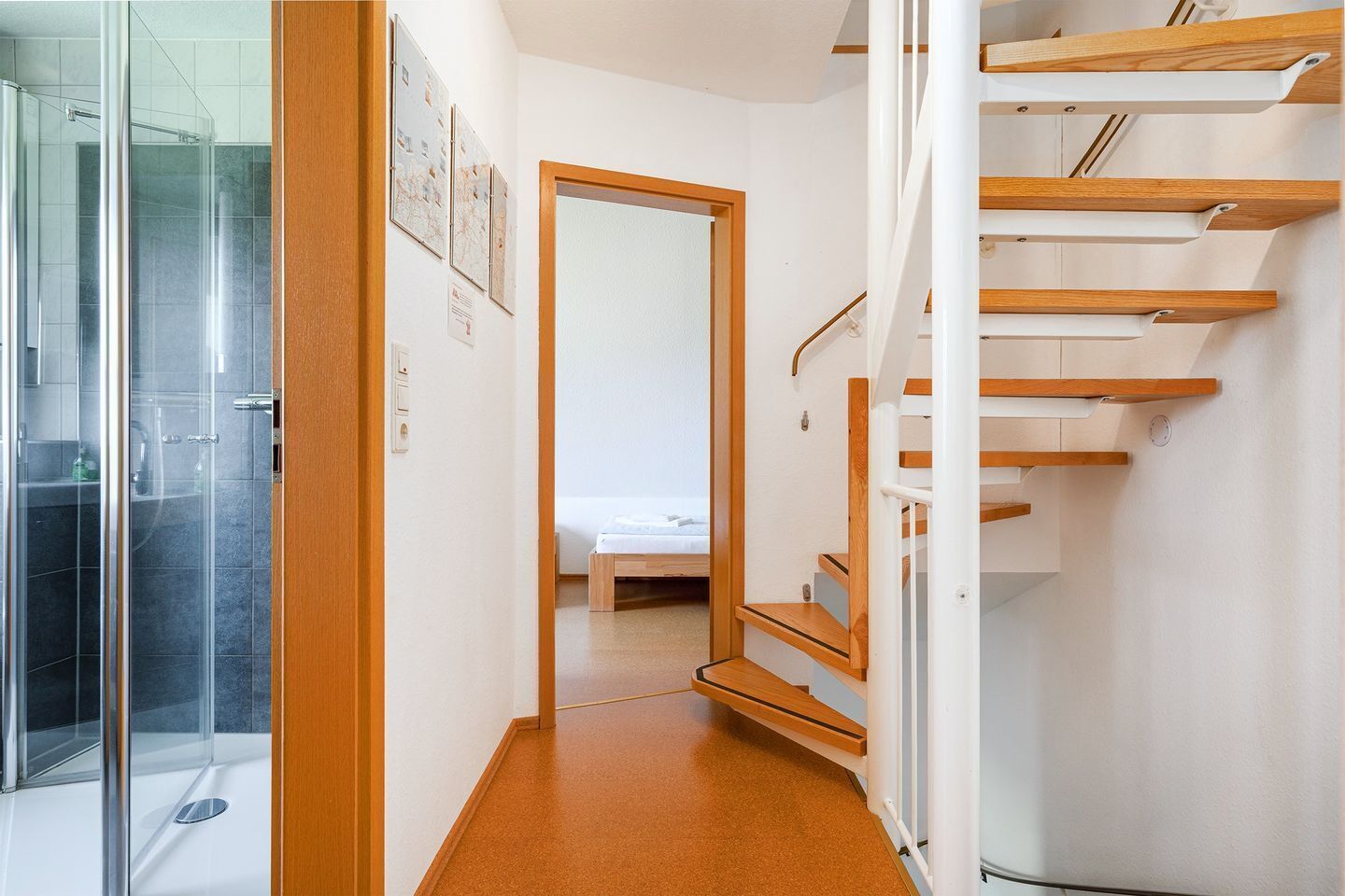 A hallway with a glass shower enclosure, a wooden staircase, and a bedroom with a bed and a nightstand.