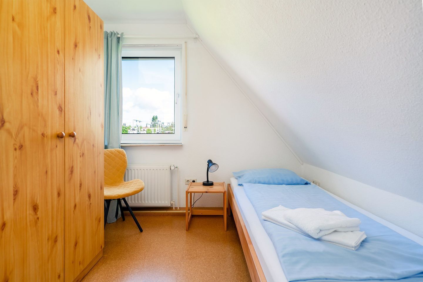 Une petite pièce propre et bien rangée, avec un lit, une chaise, un bureau et une fenêtre.