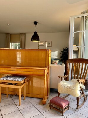 A room with a wooden piano, a small wooden table with a cushion, a rocking chair, and a lamp.