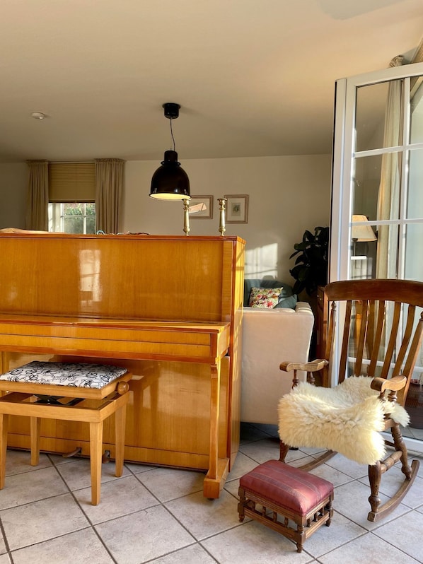 A room with a wooden piano, a small wooden table with a cushion, a rocking chair, and a lamp.