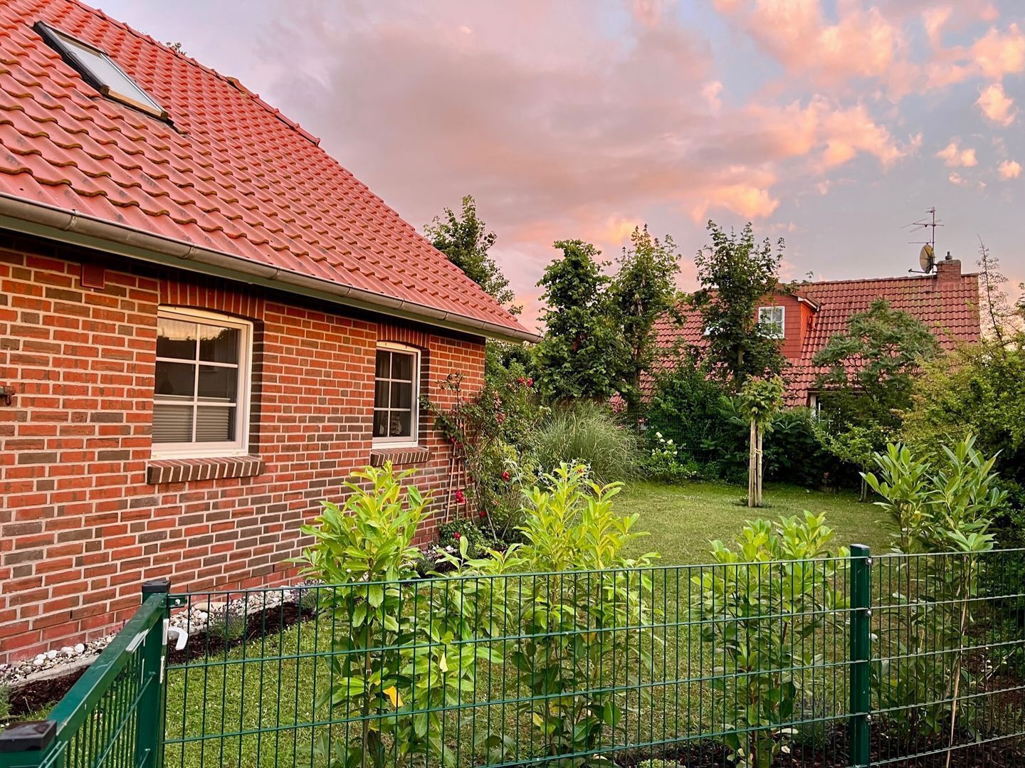 Ein Backsteinhaus mit rotem Ziegeldach, ein kleiner Garten mit Grünpflanzen und ein grüner Metallzaun.