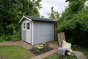 A small shed with a door and windows, surrounded by a garden with various plants and a chair with a cushion.
