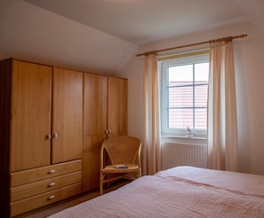 A bedroom with a wooden wardrobe, a wicker chair, a bed, and a window with curtains.