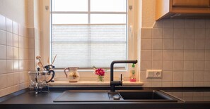 Una cocina con ventana, un lavabo, un colador, una jarra y un florero con flores.