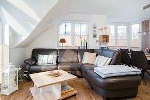 A modern living room with a brown leather sofa, a wooden coffee table, and a dining table with chairs. There are large windows providing natural light and a view of the outside. - Mühlenkieker - Cozy vacation apartment (80sqm) for 4 persons with balcony in (Krummhörn)