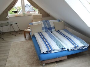 Quarto com teto inclinado, cama com roupa de cama listrada, uma cadeira, uma mesa e janela com vista para uma casa e um pássaro.