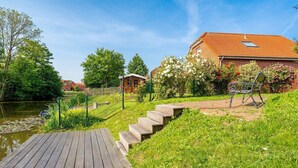 A wooden deck leading to a garden with a bench, a small building, and a house with a red roof. - Midsummer - Beautiful vacation home (100sqm) for 6 people with fireplace, garden and (Krummhörn)