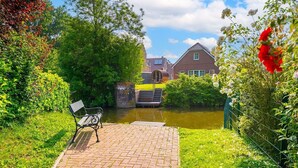 A brick pathway leading to a bench overlooking a canal, with a house and lush greenery in the background. - Midsummer - Beautiful vacation home (100sqm) for 6 people with fireplace, garden and (Krummhörn)