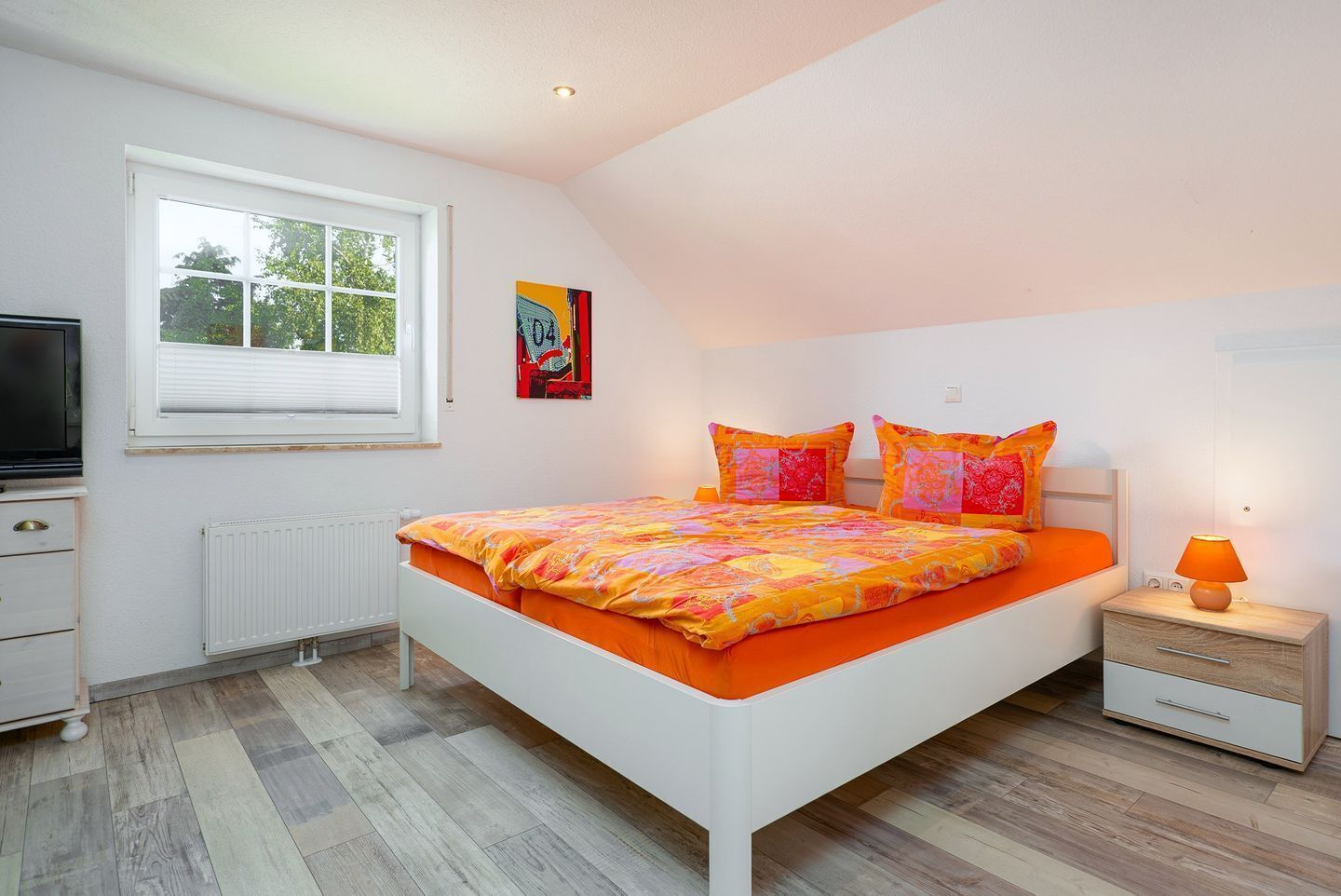 A bedroom with a white bed, orange bedding, a nightstand with a lamp, a radiator, and a window with blinds.