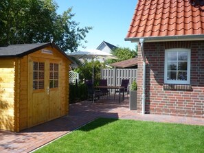 Un abri en bois et un bâtiment en briques surmonté d’un toit de tuiles rouges, une terrasse avec une table et des chaises, et une cour clôturée.